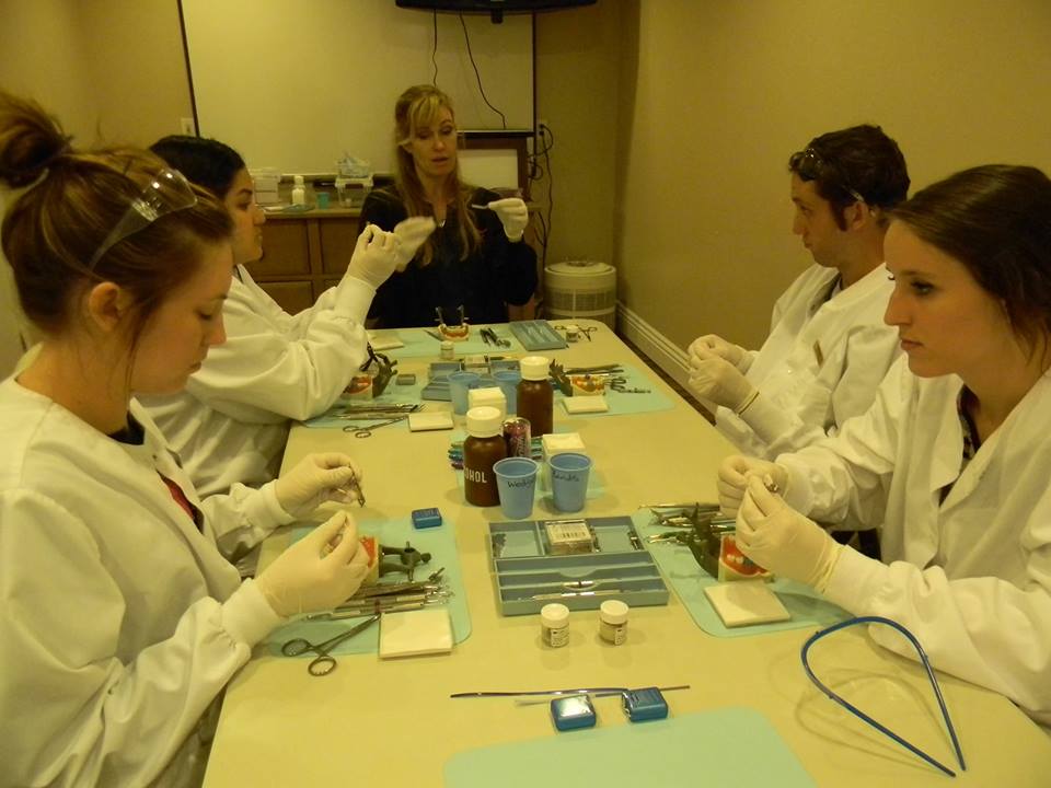 A group of people wearing white lab coats are seated at a table with various medical equipment and supplies, engaged in an activity that appears to be related to science or healthcare.