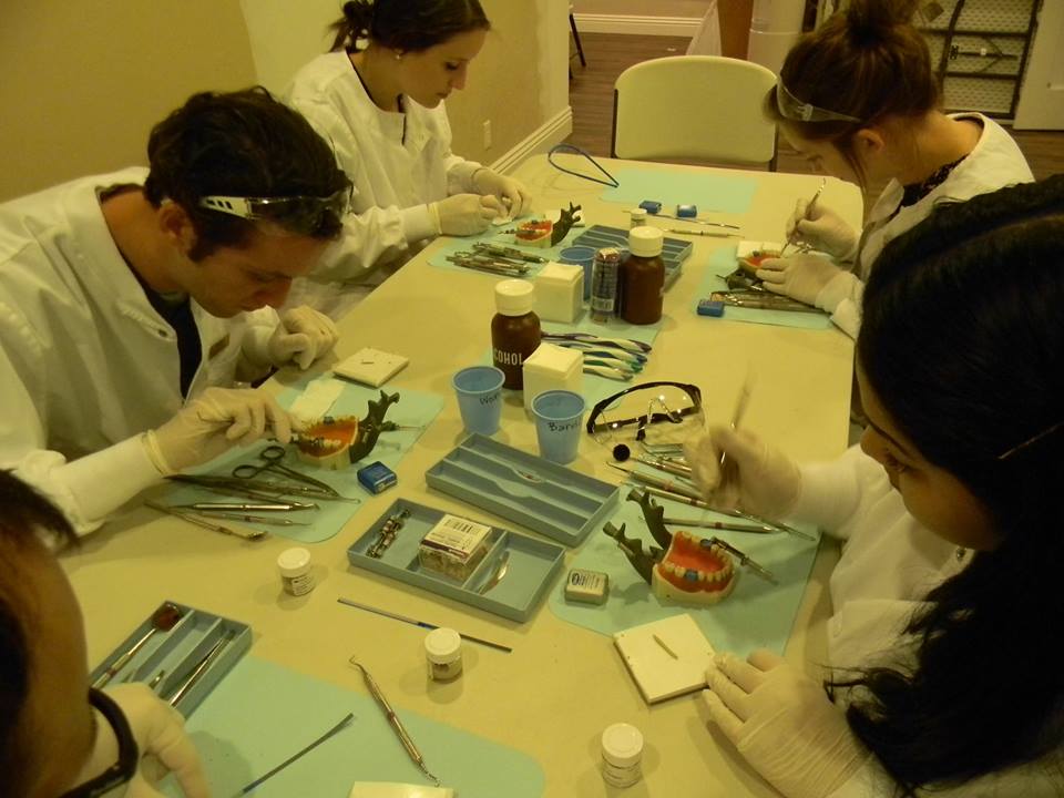 A group of people wearing lab coats are gathered around a table with various medical equipment and supplies, engaged in an activity that appears to be related to science or medicine.