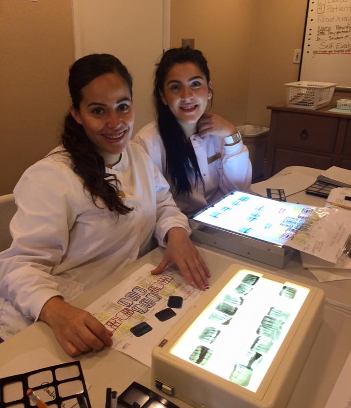 Two individuals, likely dental professionals, working with dental models at a desk with various materials and equipment, including a monitor displaying images of teeth.