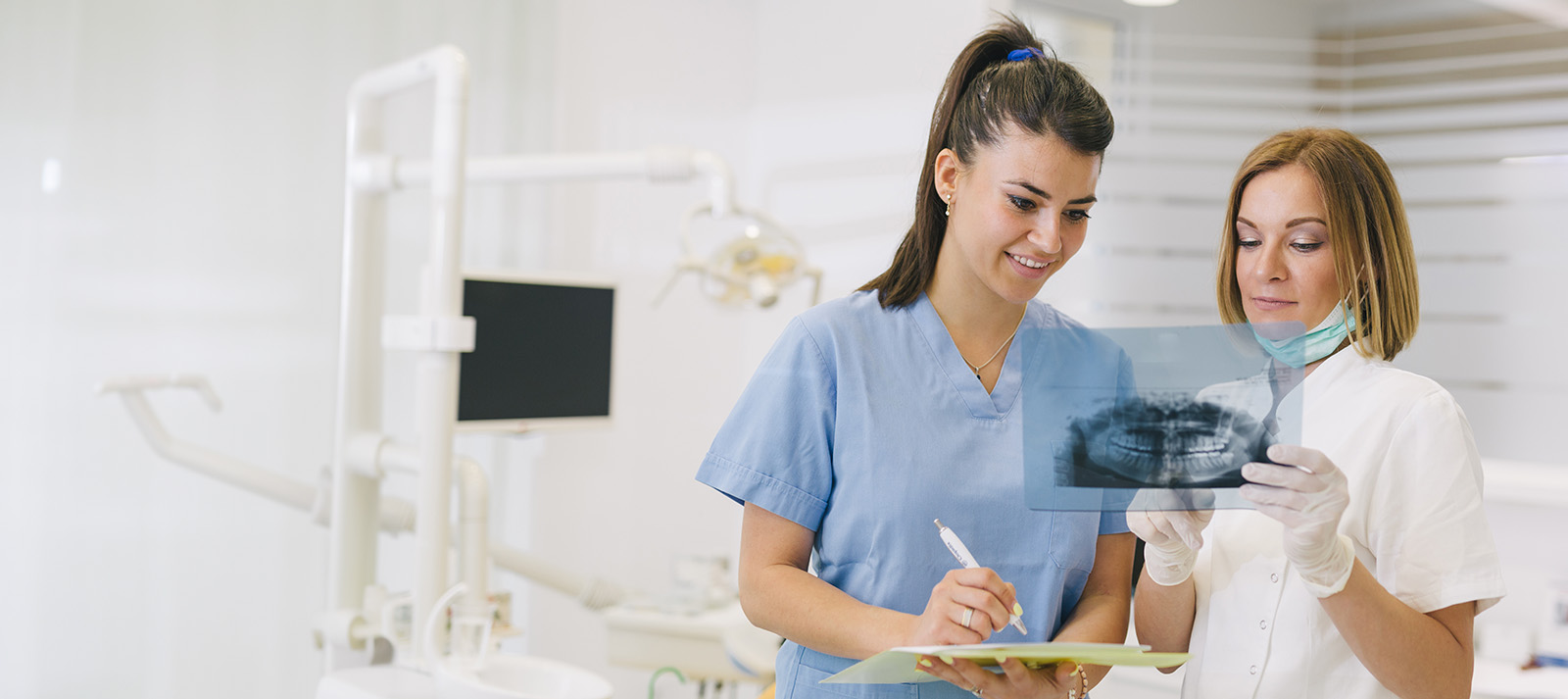 Two individuals, likely healthcare professionals, standing in front of dental equipment with a tablet displaying what appears to be medical imaging.