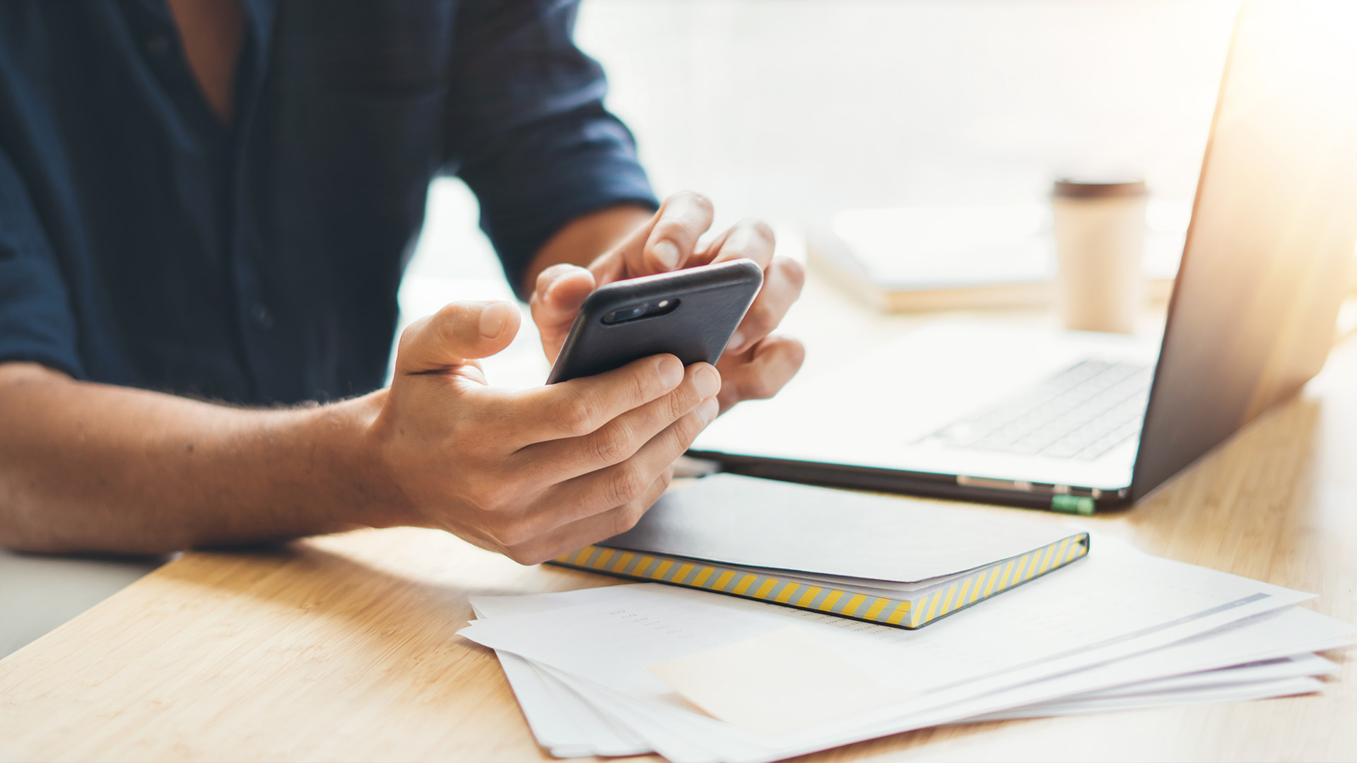 A person using a smartphone while sitting at a desk with papers and a laptop.