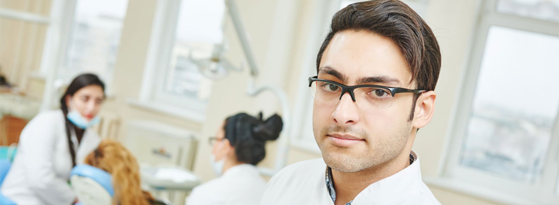 The image shows a man standing in front of a dental office setting with dental professionals seated behind him, wearing lab coats and working with equipment.