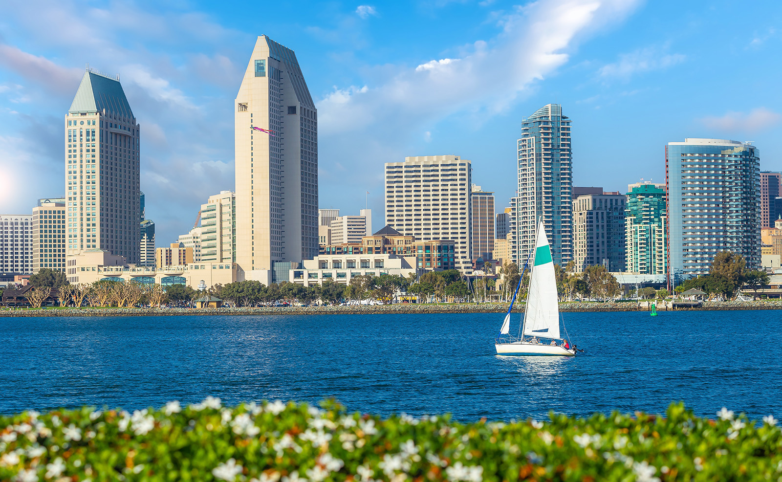 An urban waterfront scene with a sailboat on the river, city skyline, and a clear blue sky.