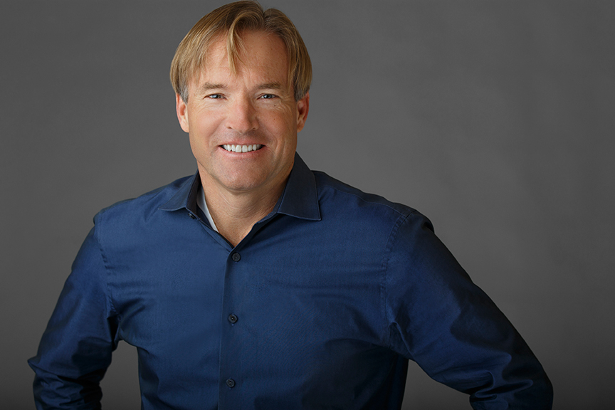 The image features a man standing against a neutral background, wearing a dark shirt with rolled-up sleeves and leaning slightly forward with one hand on his hip.